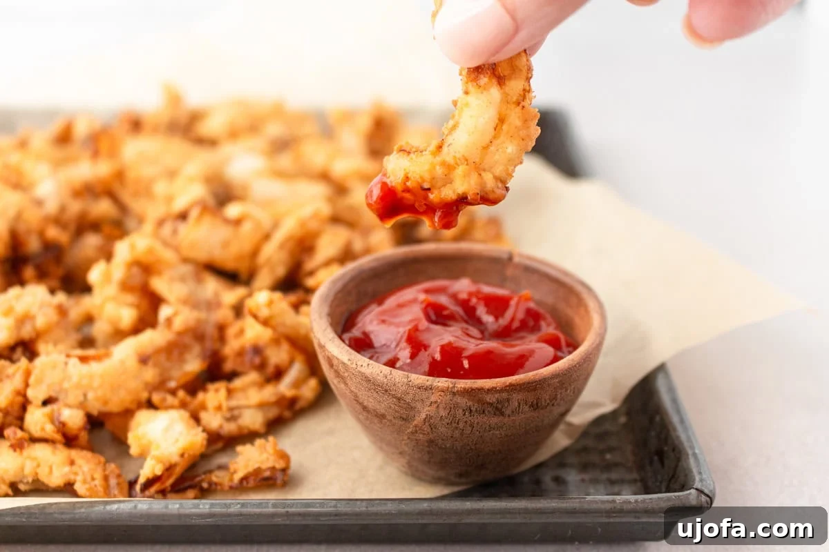 A tray of golden-brown gluten-free fried onions, with one piece being dipped into a small bowl of tomato sauce, highlighting their versatility.