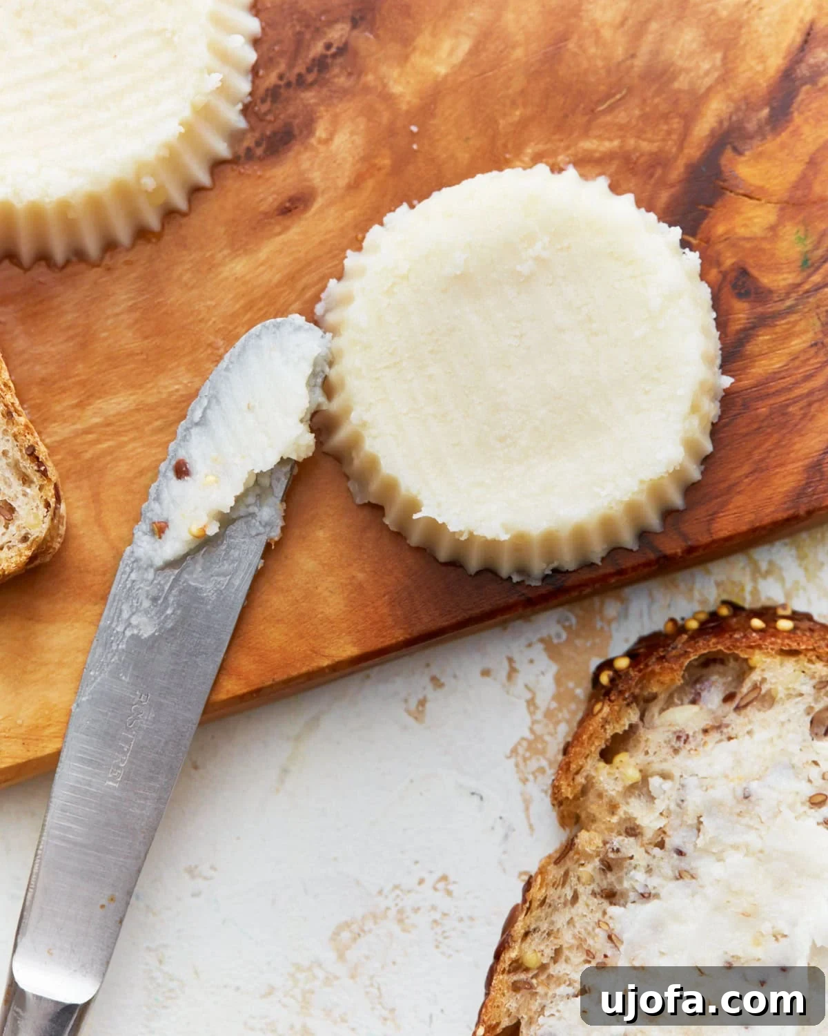 A golden block of homemade vegan butter sits on a wooden cutting board, accompanied by a butter knife and a piece of buttered toast, highlighting its creamy texture.