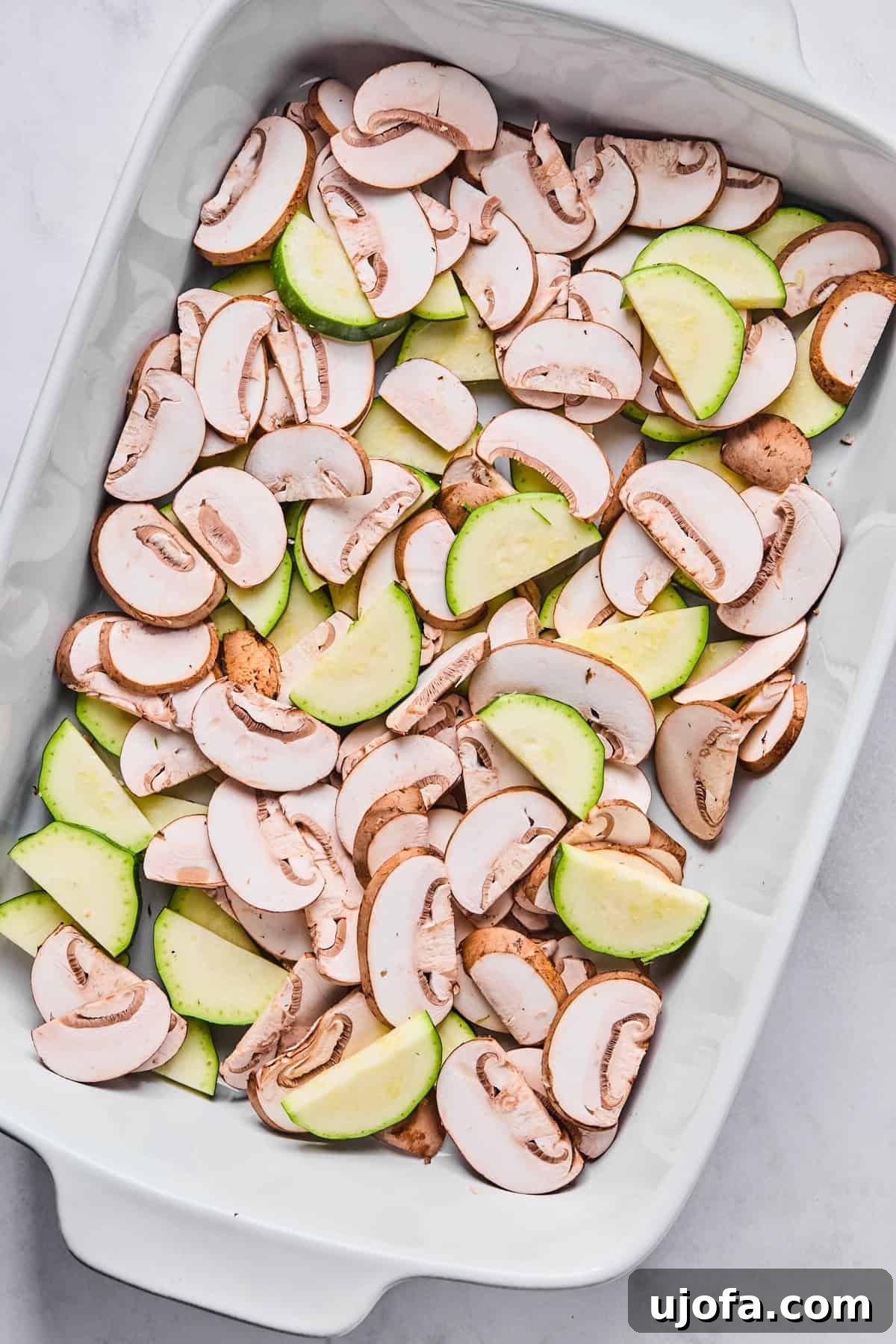 Sliced mushrooms and zucchini spread out evenly in the bottom of a 9x13-inch casserole dish, ready for baking.