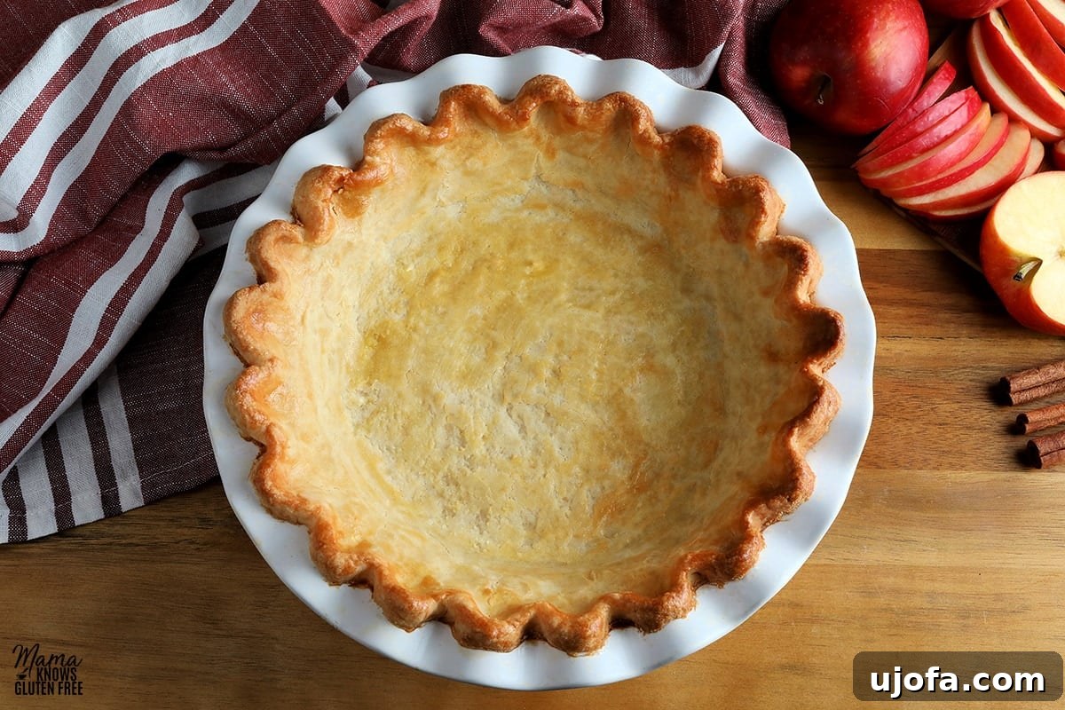 A perfectly golden, baked gluten-free pie crust filled with sliced apples, ready for a top crust or crumble, sits on a wooden surface with a rustic red kitchen towel draped in the background, highlighting the quality of the crust.