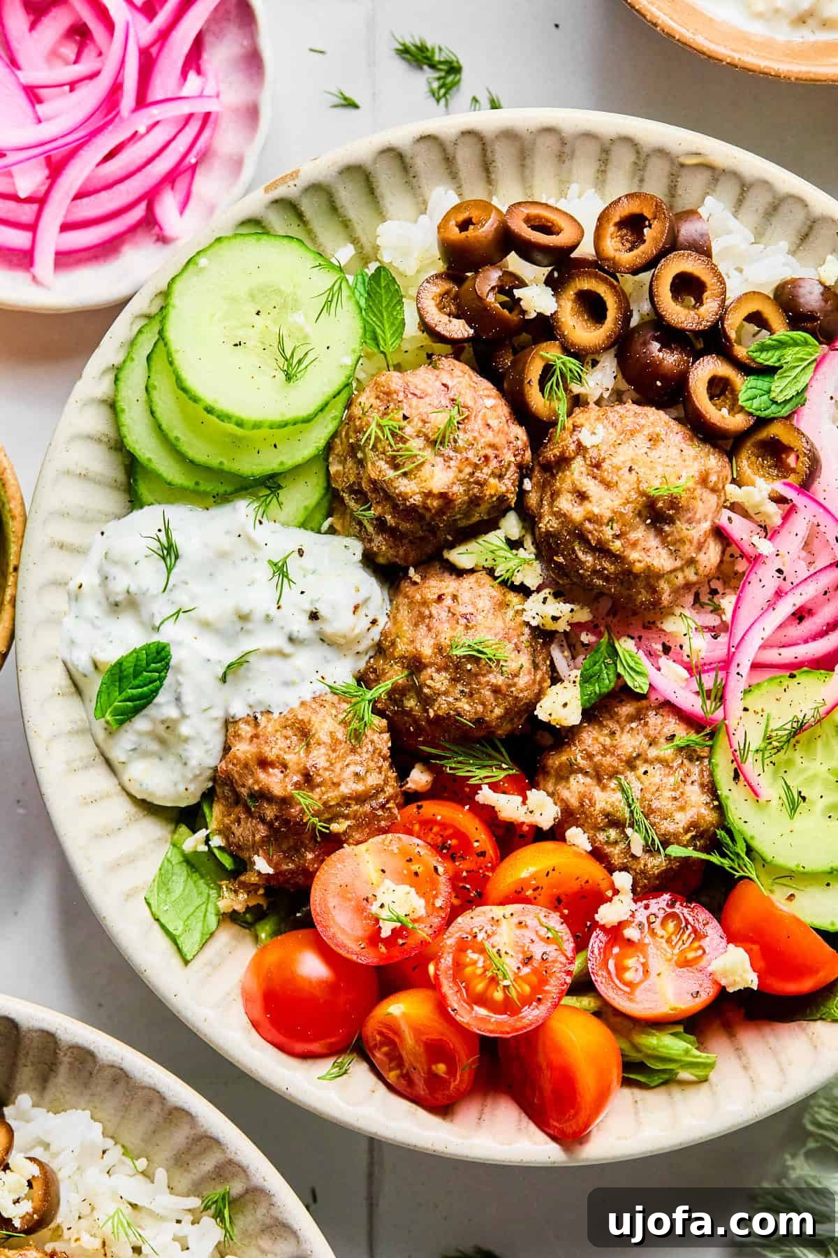 Close-up of Greek lamb meatballs served over rice, surrounded by sliced cucumber, black olives, red onion, cherry tomatoes, fresh herbs, and crumbled feta cheese, with lemon dill yogurt sauce on the side.