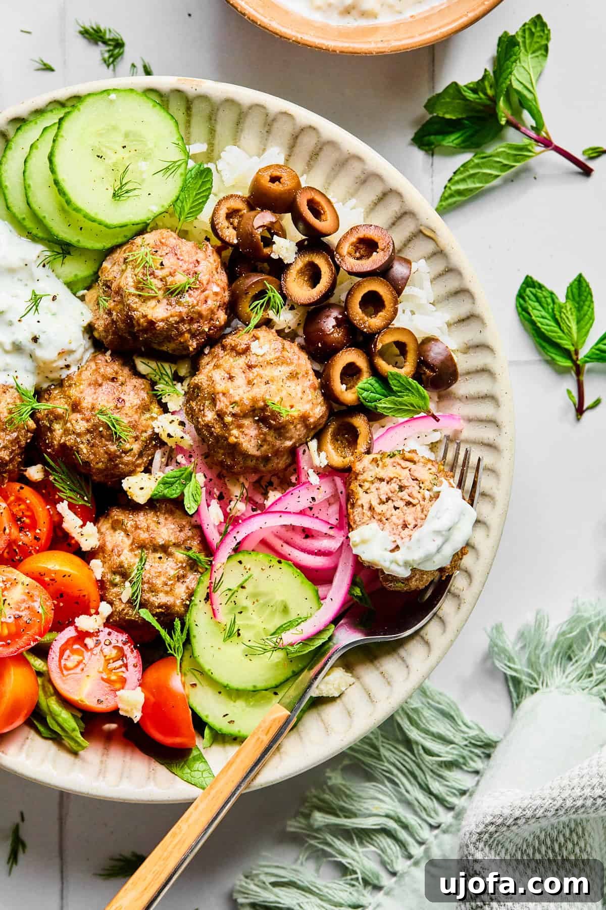 Greek lamb meatballs served over rice, surrounded by sliced cucumber, black olives, red onion, cherry tomatoes, fresh herbs, and crumbled feta cheese, with lemon dill yogurt sauce.