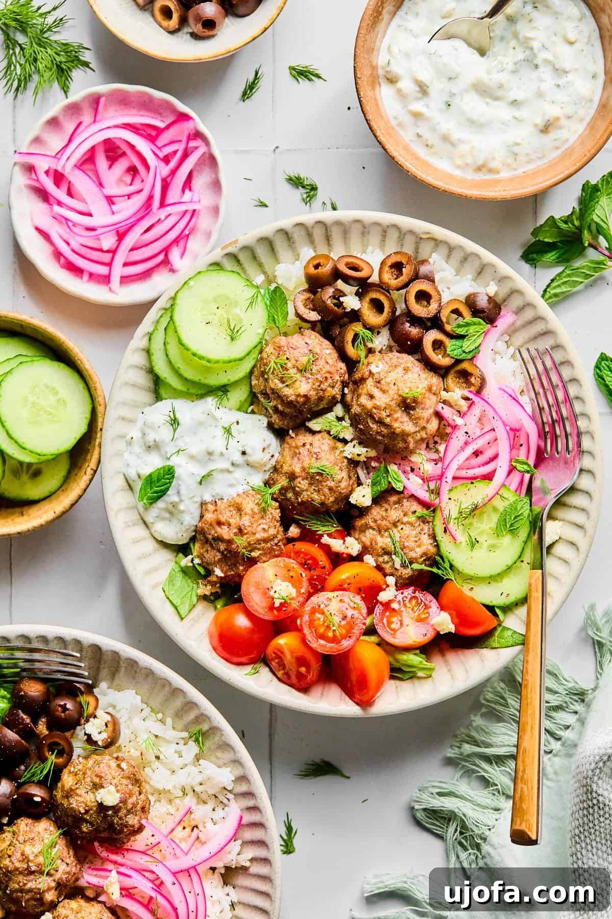 Greek lamb meatballs served over rice, surrounded by sliced cucumber, black olives, red onion, cherry tomatoes, fresh herbs, and crumbled feta cheese, with lemon dill yogurt sauce.