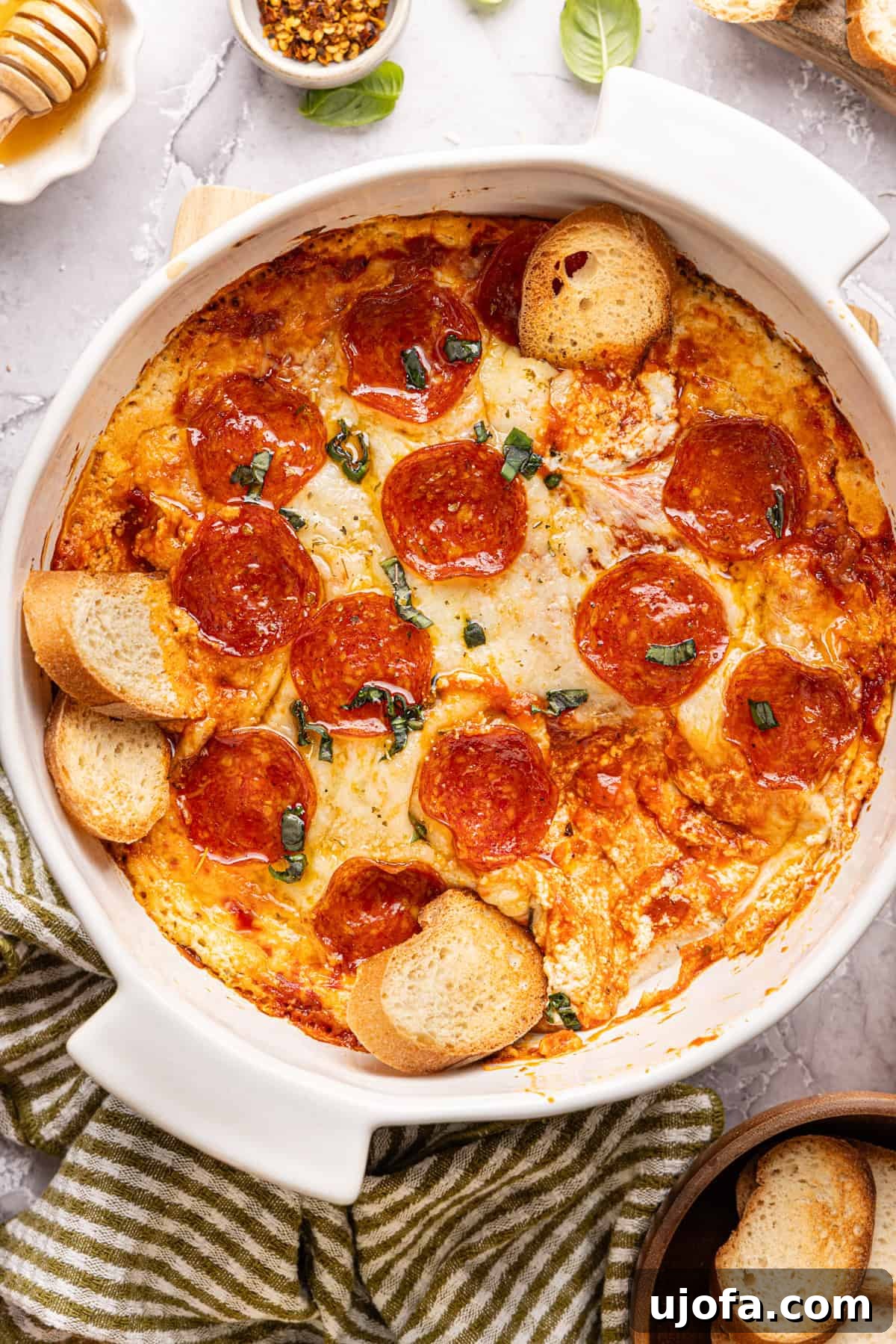 Ultimate Creamy Protein Pizza Dip 2 A perfectly baked high protein pizza dip in a rustic round white dish, garnished generously with vibrant pepperoni, luscious melted cheese, a delicate drizzle of hot honey, and fresh, aromatic basil leaves. A few small, crusty bread slices are strategically placed within the dip, inviting indulgence.
