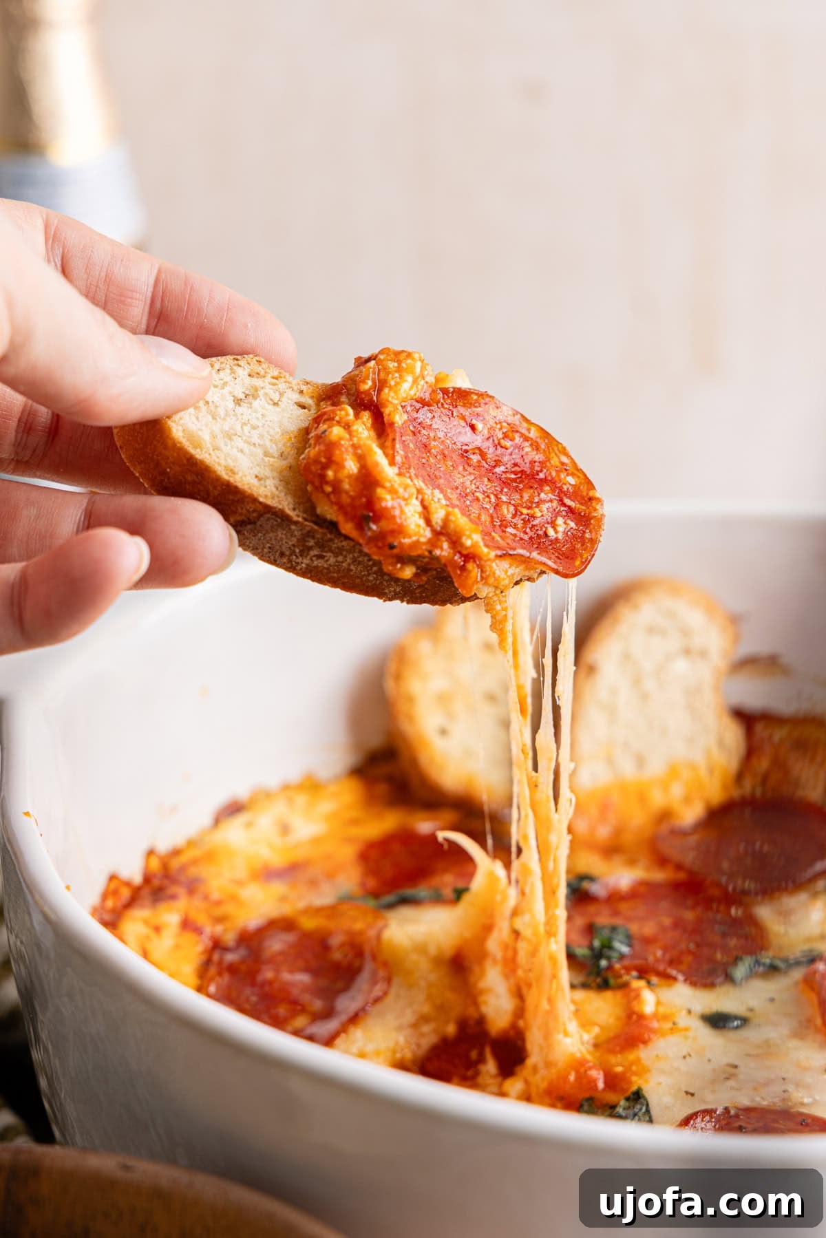 Ultimate Creamy Protein Pizza Dip 13 A hand holds a golden slice of perfectly toasted bread, generously topped with a rich scoop of high protein pizza dip, poised above the communal dish still abundant with the remaining irresistible dip, inviting everyone to partake.