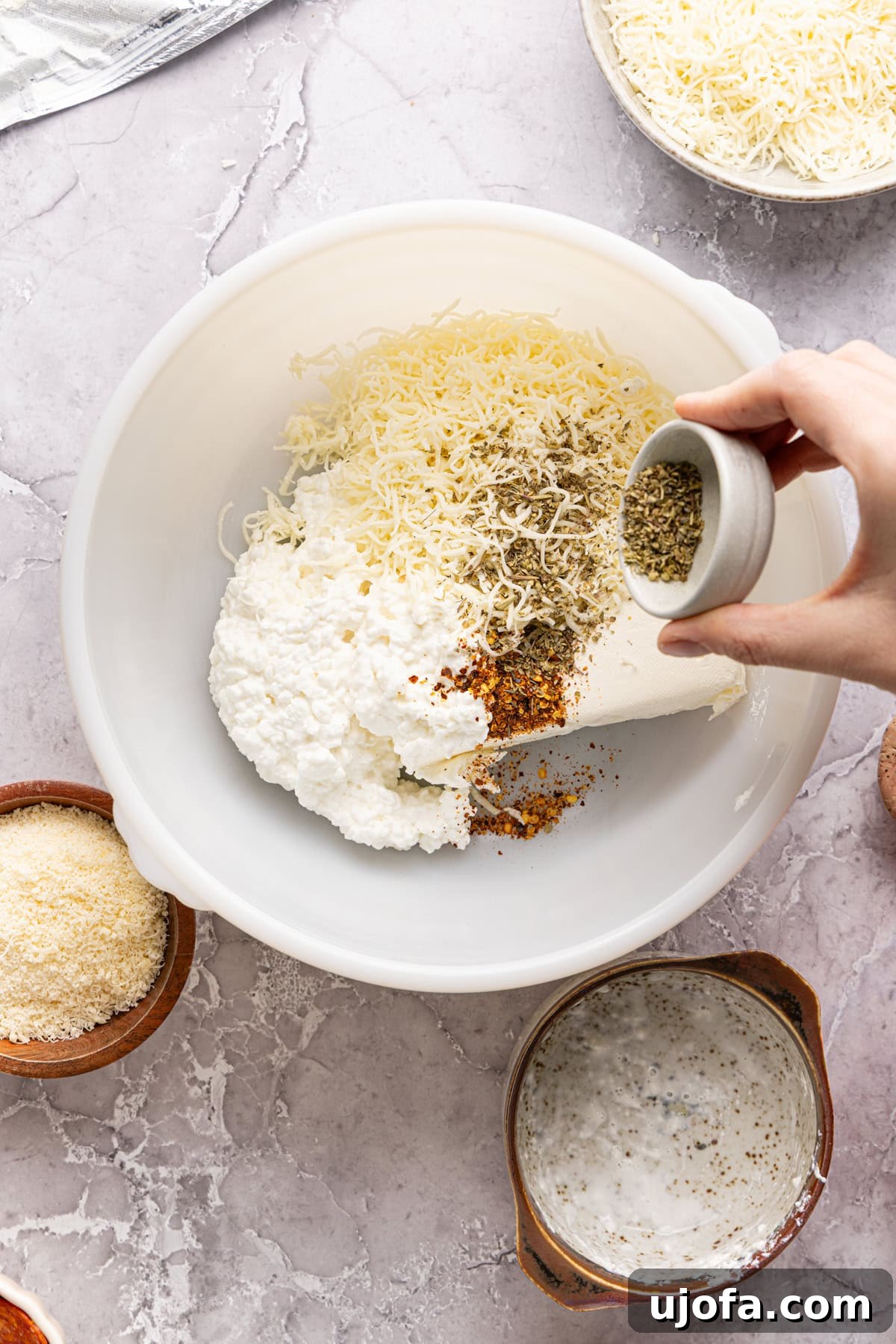 Ultimate Creamy Protein Pizza Dip 4 A pristine white mixing bowl filled with a creamy, blended mixture of softened cream cheese, cottage cheese, shredded mozzarella, grated parmesan, and various aromatic seasonings. A handheld electric mixer rests gently inside the bowl, indicating the blending process is either underway or just completed.