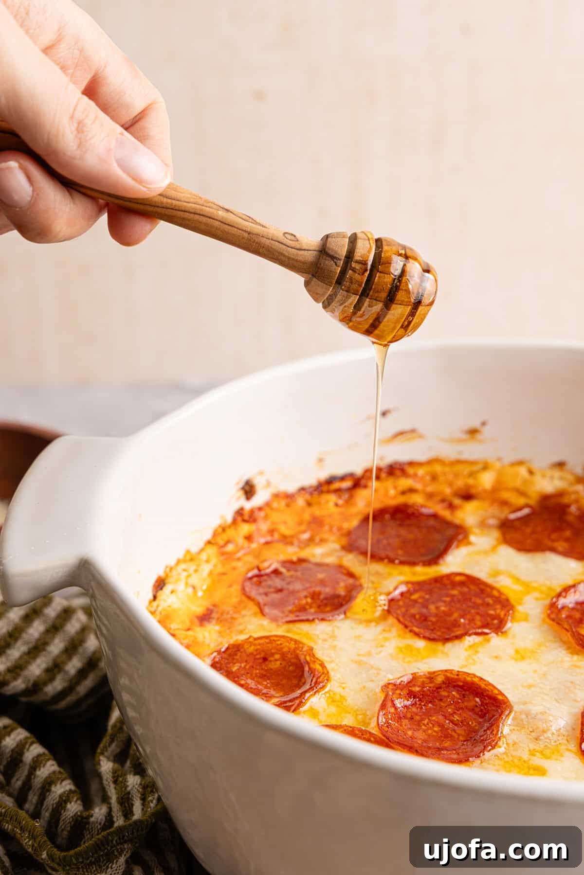 Ultimate Creamy Protein Pizza Dip 10 A human hand gracefully drizzling shimmering hot honey over the freshly baked, bubbly high protein pizza dip, adding a sophisticated touch of sweet heat and glistening sheen.