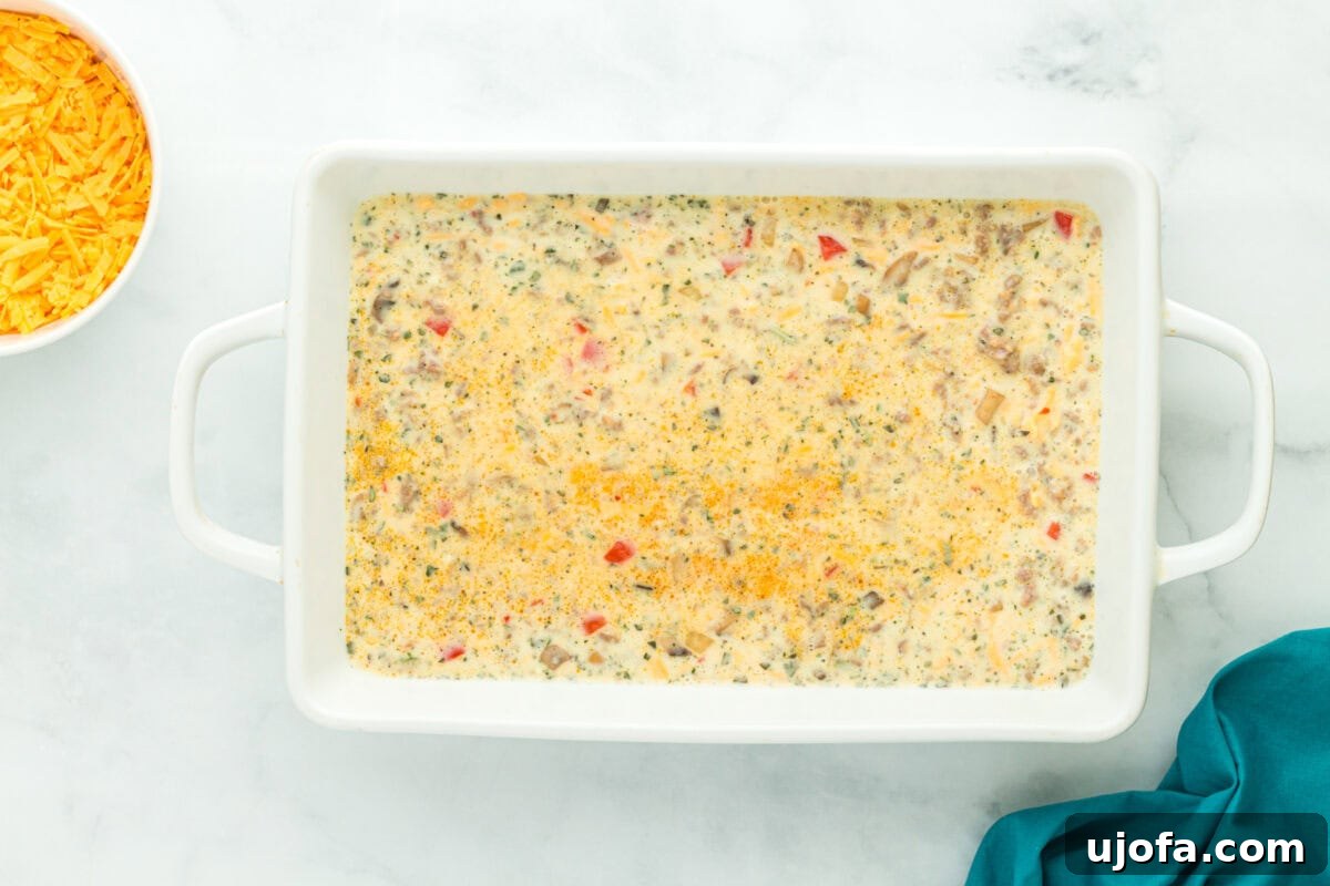 Yuletide Sausage Strata 12 An inviting overhead view of an uncooked breakfast casserole, brimming with savory sausage, colorful vegetables, and creamy eggs, generously topped with shredded cheese in a white baking dish, prepared and ready to be baked to golden perfection.
