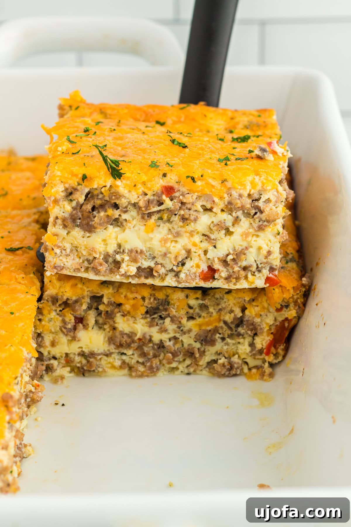 Yuletide Sausage Strata 4 A spatula gently lifts a perfectly baked piece of the savory breakfast sausage casserole from a white baking pan, showcasing its golden crust and fluffy interior, ready for serving.