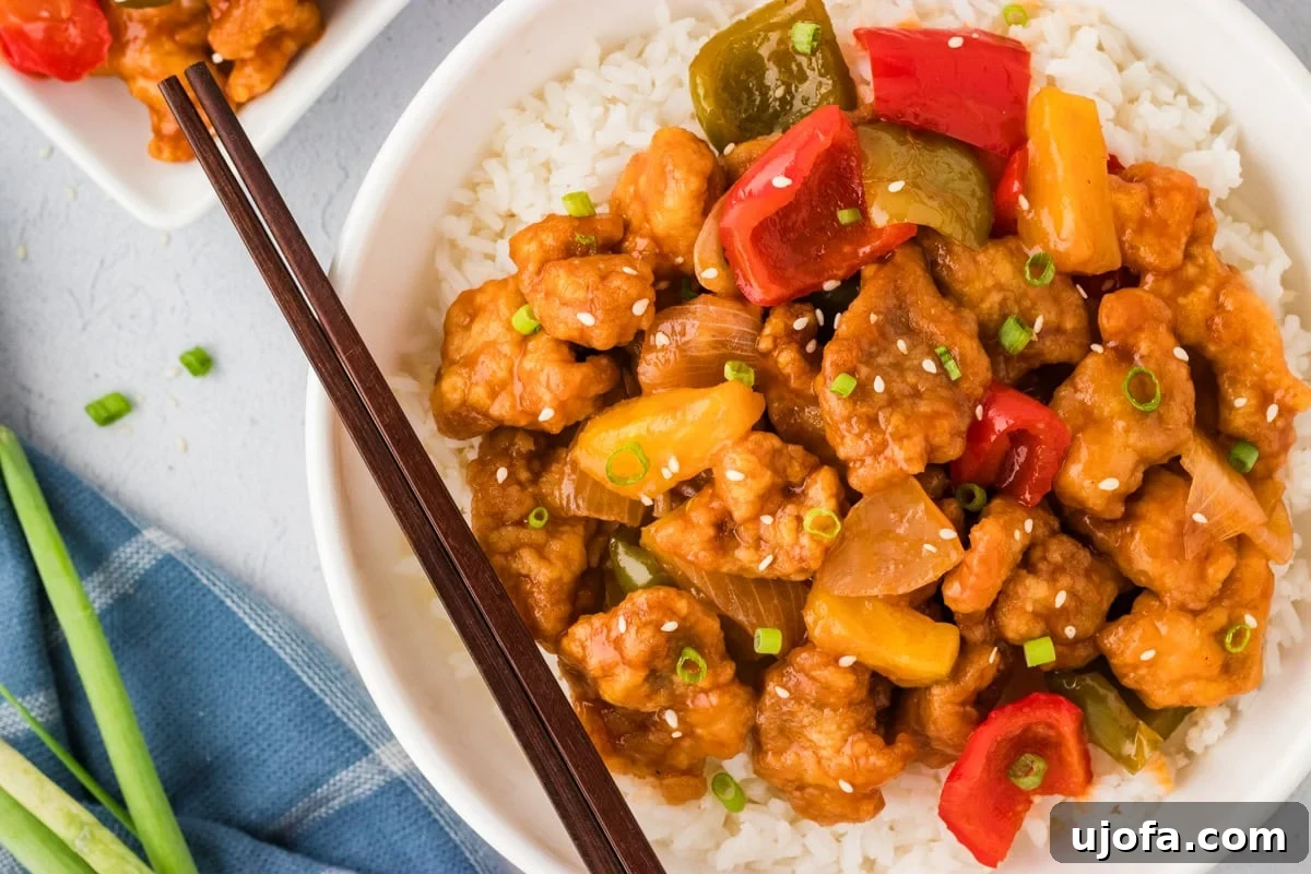 Overhead view of a perfectly plated gluten-free sweet and sour chicken with stir-fry vegetables over rice in a white bowl, garnished with sesame seeds and green onions, with chopsticks ready.