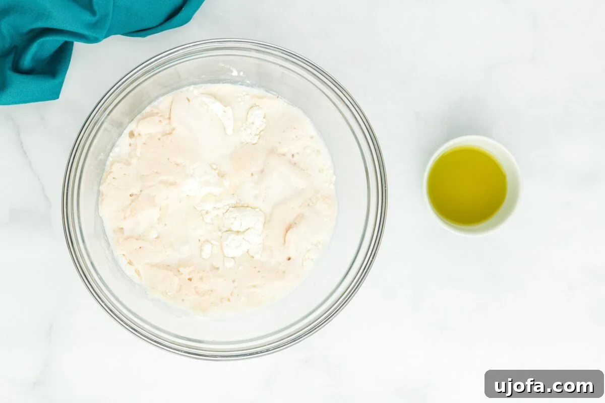 The combined gluten-free bao bun dough, wet and dry ingredients, forming a cohesive mass within a glass mixing bowl, with a small bowl of olive oil on the side.