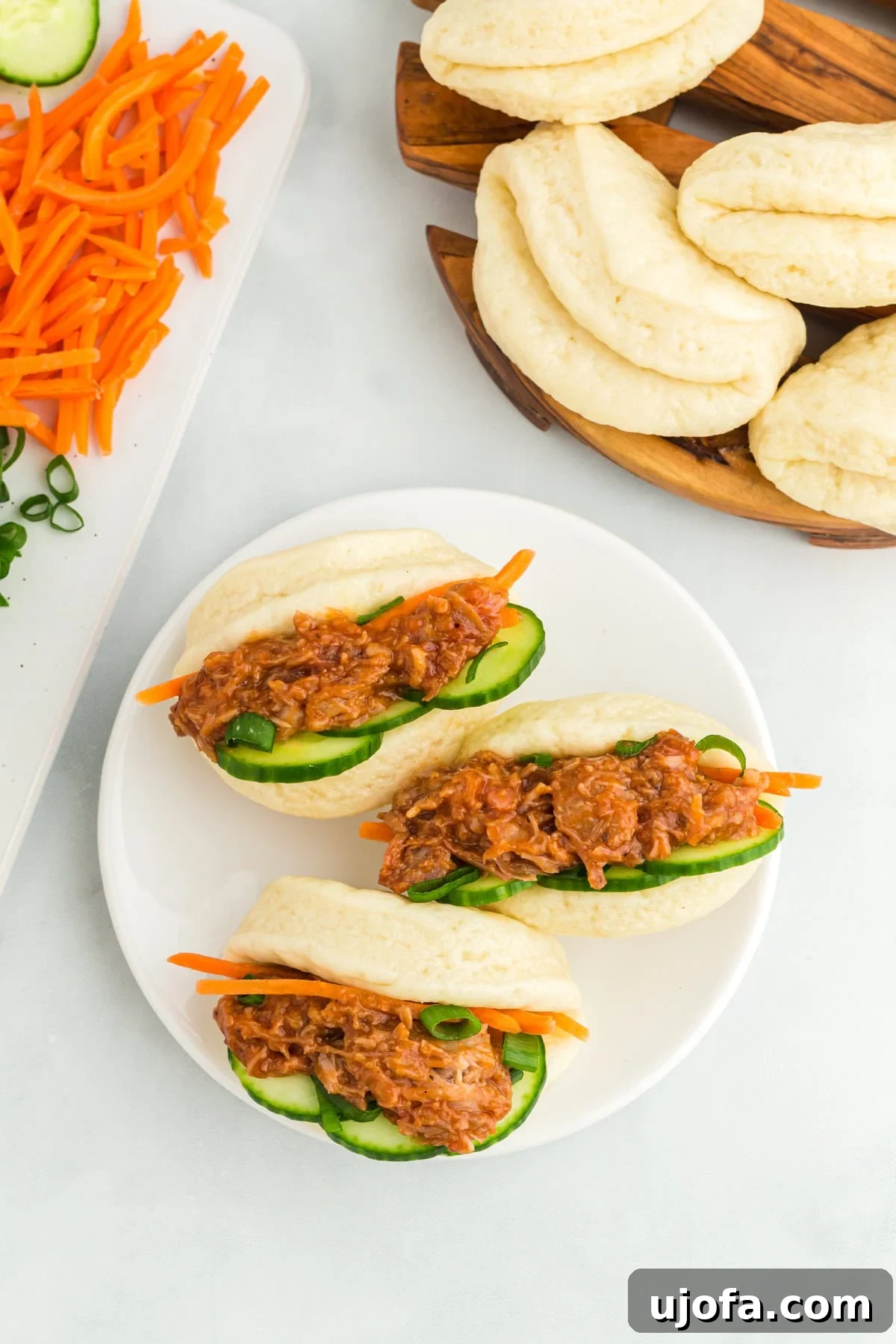 Three stuffed gluten-free bao buns, generously filled with savory meat, crisp cucumbers, and shredded carrots, elegantly arranged on a white plate.