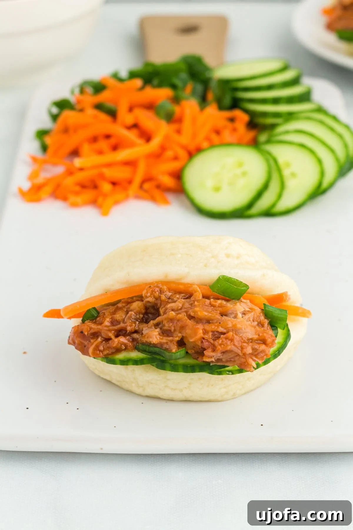 A close-up shot of a single gluten-free bao bun, artfully filled with tender meat, fresh cucumbers, crisp carrots, and vibrant green onions, resting on a white board.