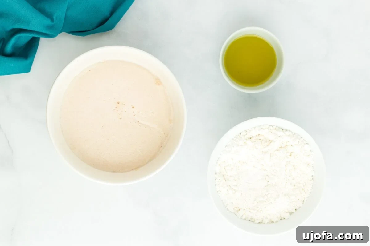 The activated yeast and liquid mixture in a white bowl, ready to be added to a larger bowl of dry flour ingredients, with a small bowl of oil nearby.
