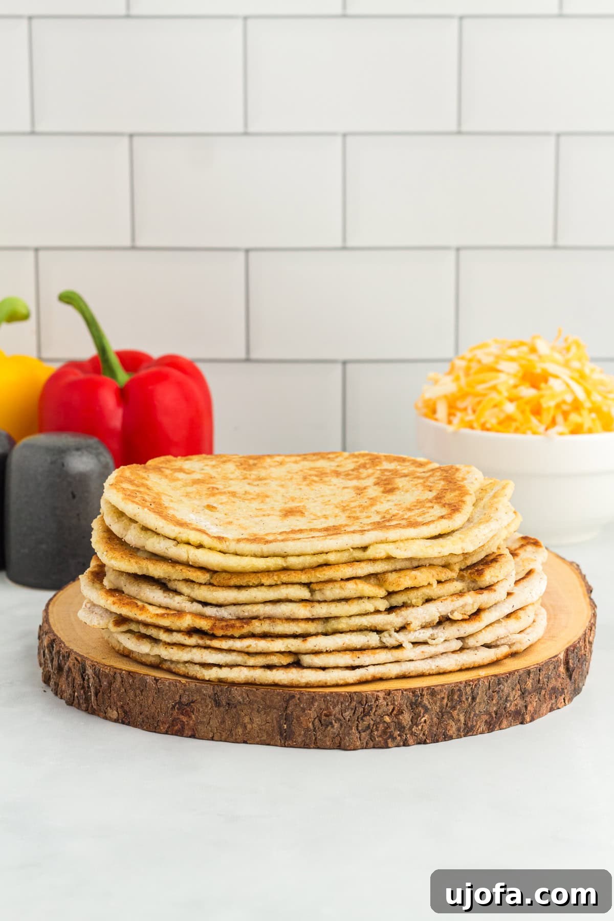 A stack of golden brown coconut flour tortillas on a wooden board, with fresh bell peppers and shredded cheese in the background, ready for a delicious meal.
