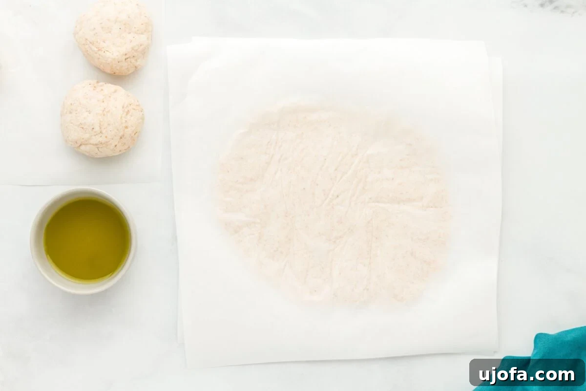 A thin, round coconut flour tortilla dough rolled flat between two sheets of parchment paper, next to a bowl of olive oil.