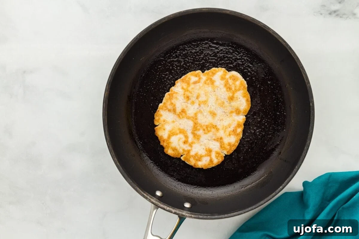 A golden-brown coconut flour tortilla cooking in a black, olive oil-greased skillet, with a spatula visible, ready to be flipped.