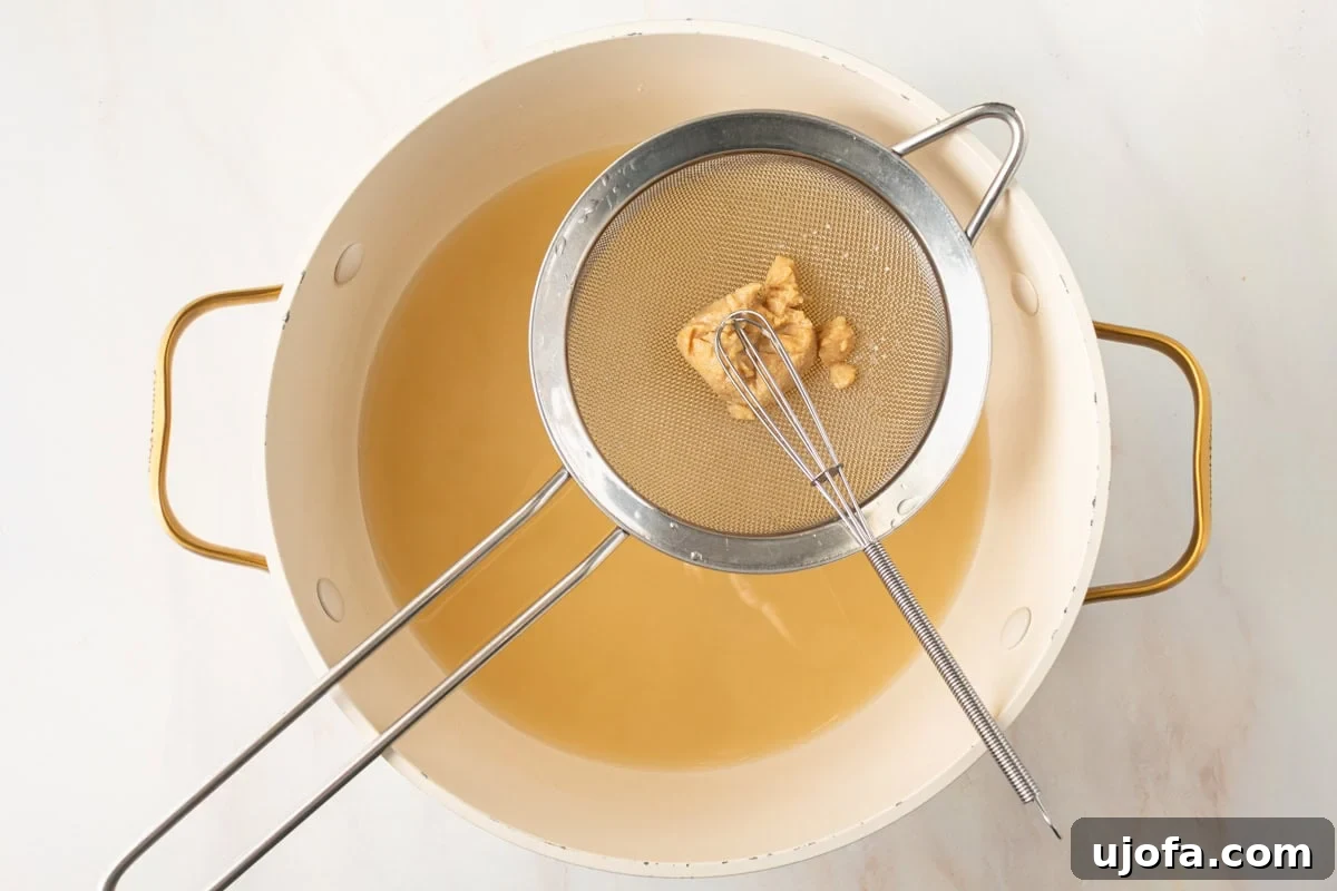 a small sieve with white miso paste being whisked into gluten-free miso soup in a large pot