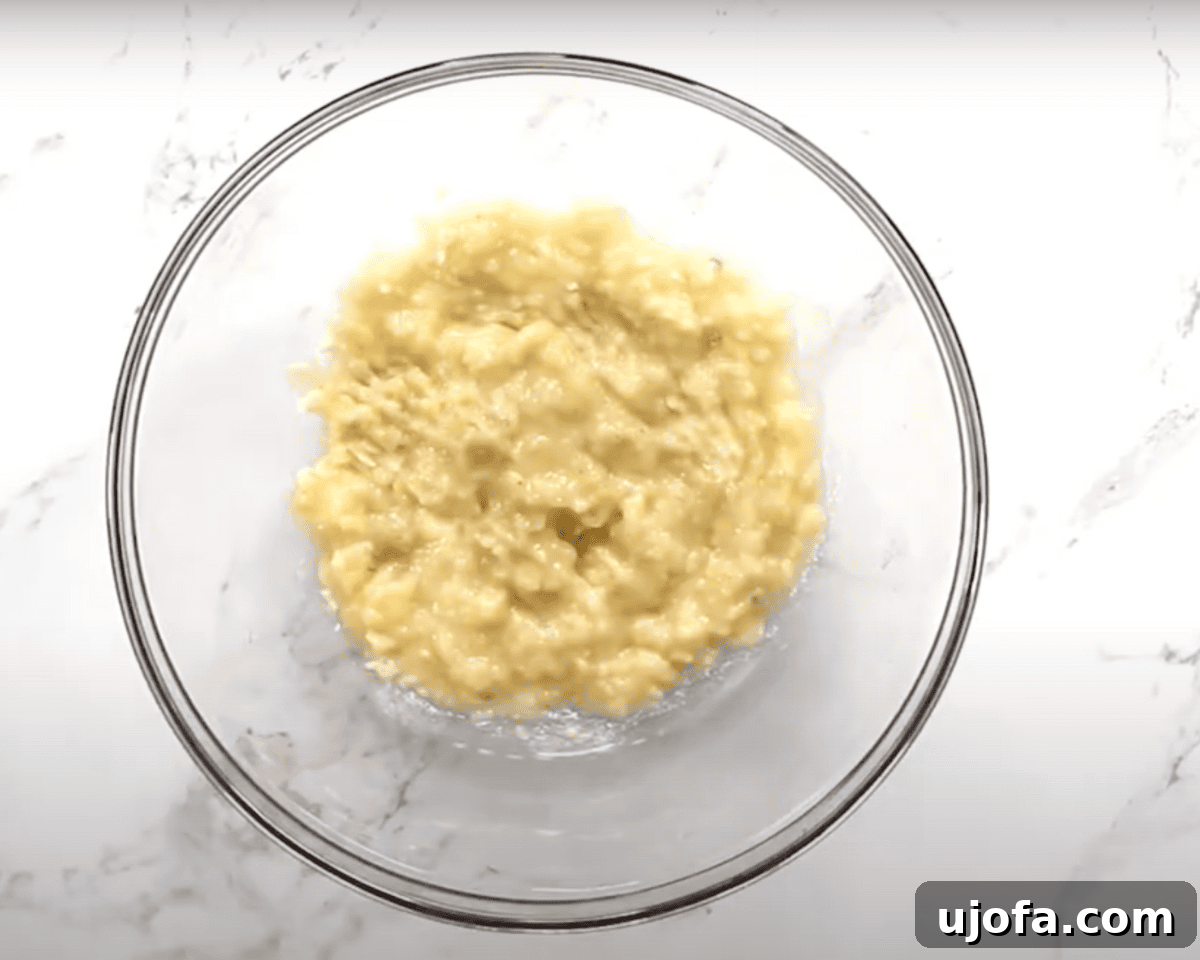 A clear glass mixing bowl filled with mashed bananas on a white marble countertop, ready for baking.