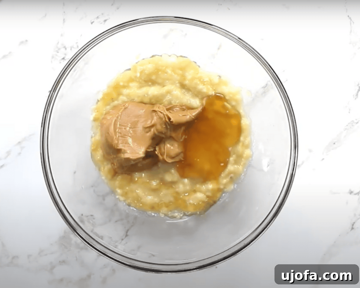 A glass bowl containing mashed bananas with scoops of peanut butter and a drizzle of honey on top, before mixing.