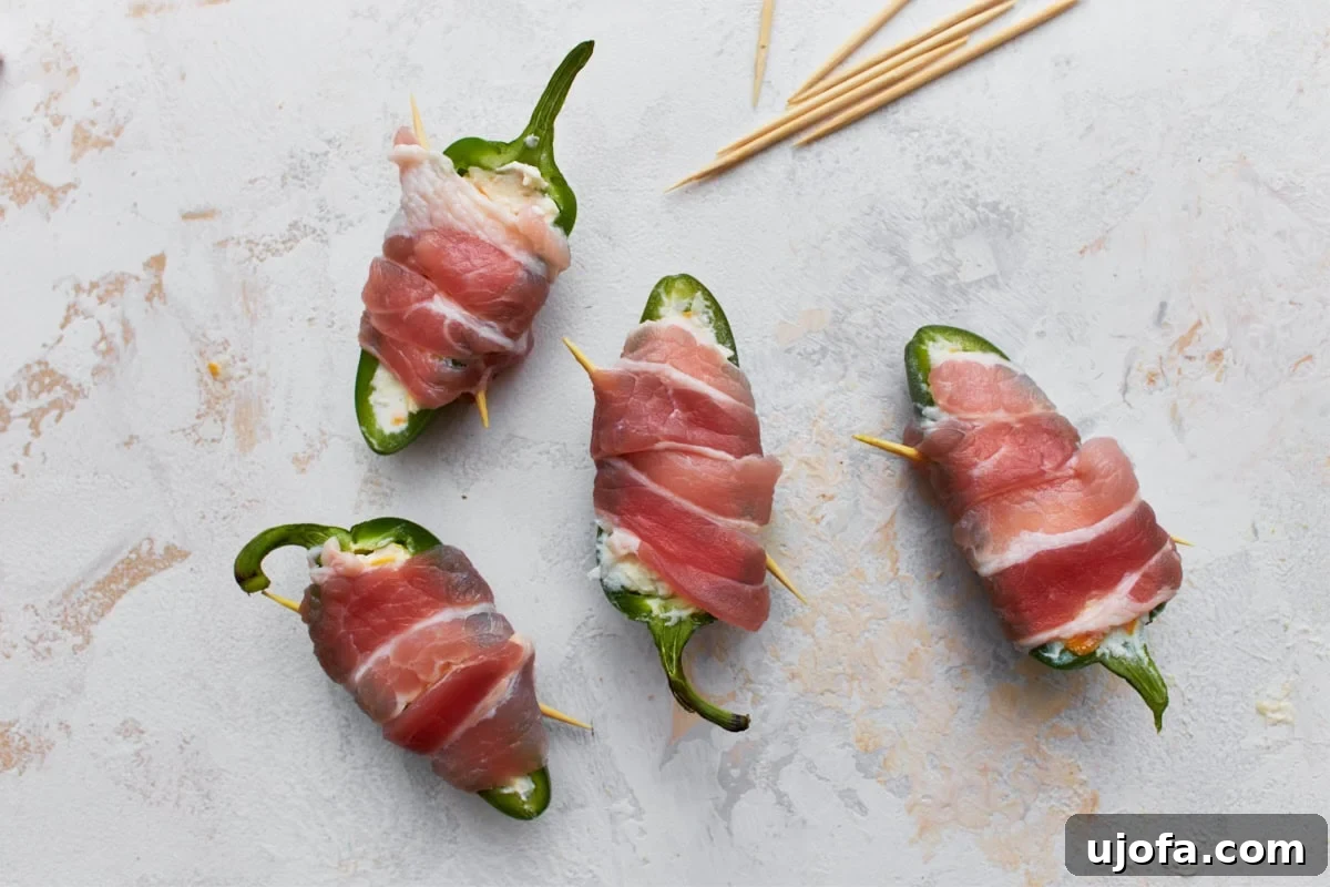 Four beautifully stuffed jalapeño halves, each meticulously wrapped with a crisp slice of bacon and secured with a toothpick, ready for baking.