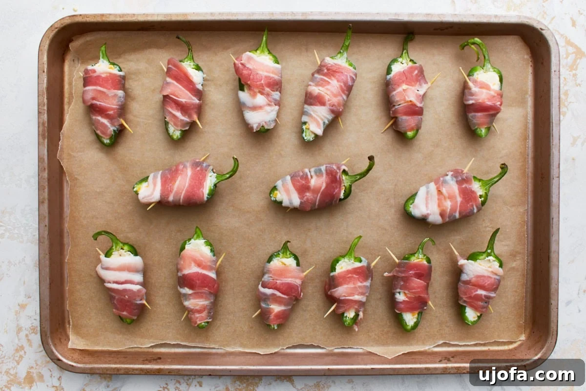 A baking sheet lined with parchment paper, holding an array of bacon-wrapped and cheese-filled jalapeño poppers, meticulously arranged and ready for the oven.