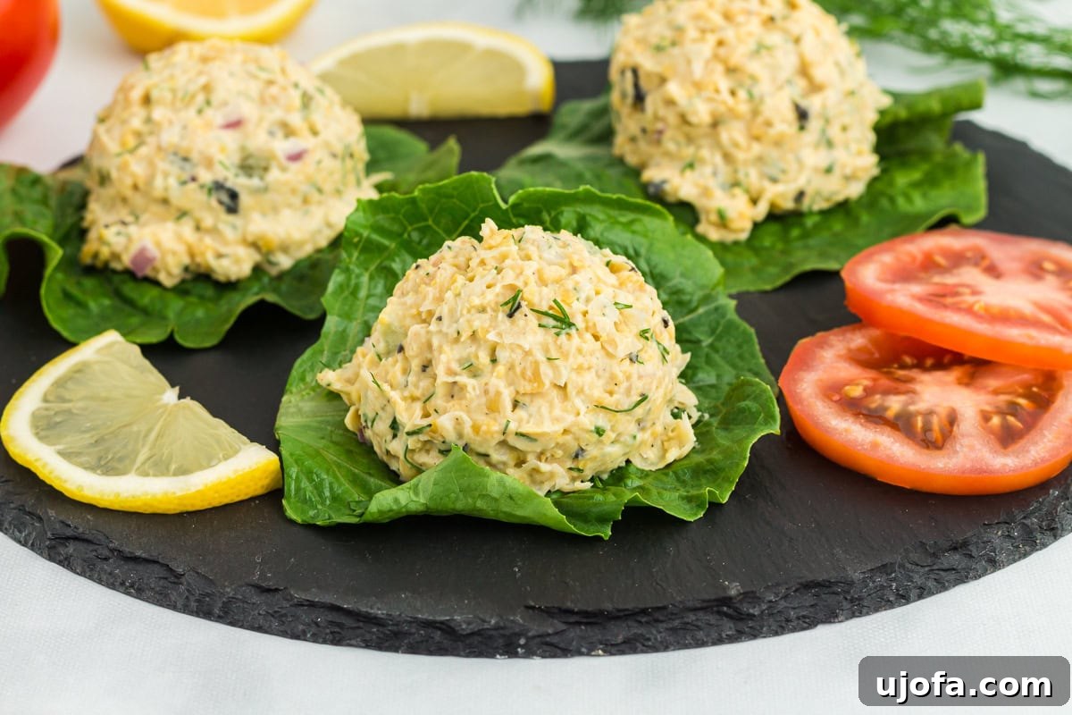 Vegan chickpea 'tuna' salad scoops served on fresh lettuce leaves, garnished with dill, lemon wedges, and tomato slices on a black slate platter.