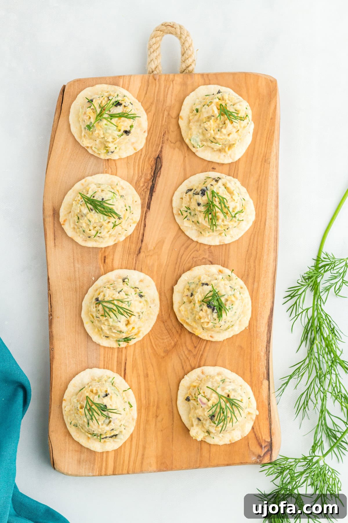 A wooden serving board with eight crackers topped with vegan chickpea tuna salad, garnished with fresh dill. The background includes a sprig of dill and a teal napkin on a white surface