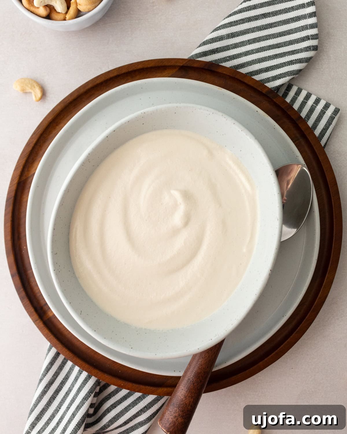 A bowl of cashew cream with a silky smooth surface, placed on a wooden tray and surrounded by cashew nuts and a striped napkin.