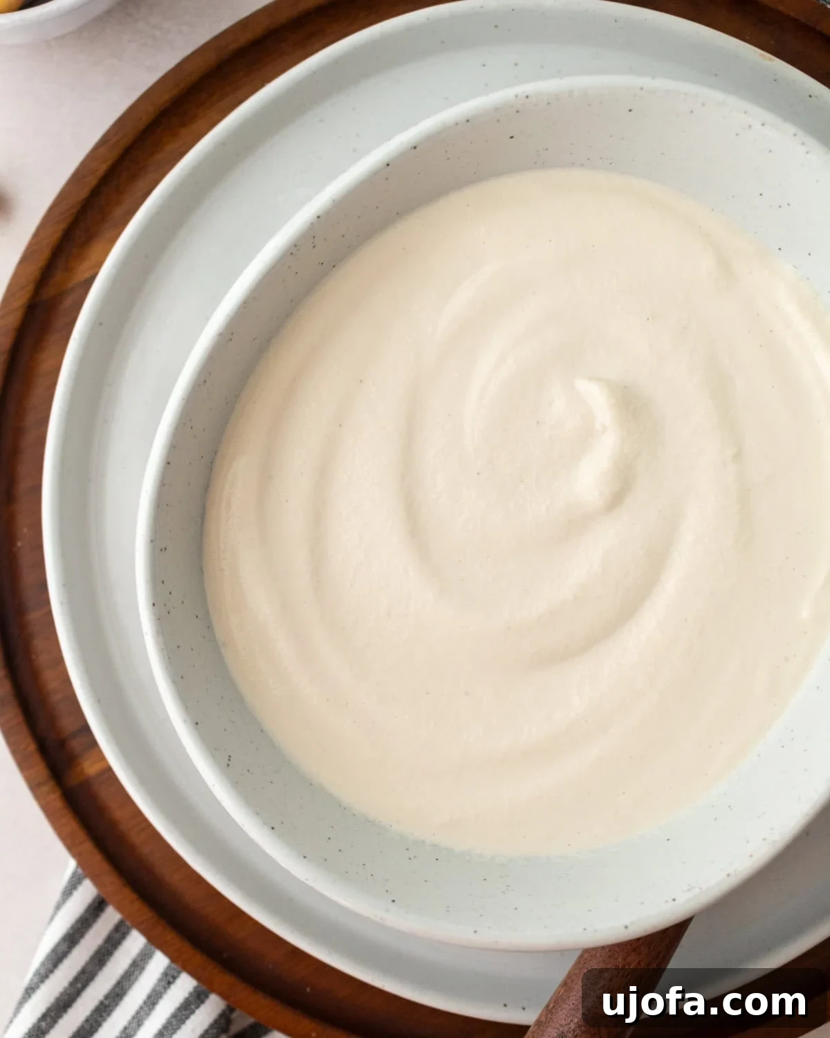 A closeup of homemade cashew cream in a white bowl. The bowl is resting on a white plate on top of a circular wooden cutting board with a spoon resting on the plate
