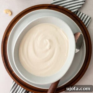 A bowl of cashew cream with a silky smooth surface, placed on a wooden tray and surrounded by cashew nuts and a striped napkin.