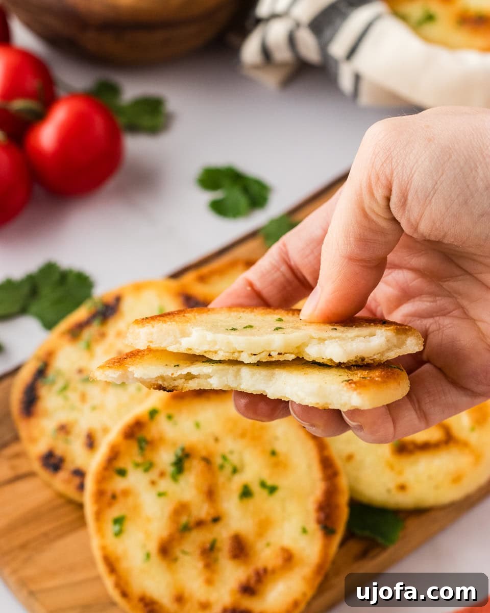 Crafting Perfect Arepas 14 A close-up of a hand holding an arepa, split open to reveal a fluffy interior. Other arepas rest on a wooden board in the background, garnished with parsley and surrounded by fresh tomatoes and cilantro leaves.