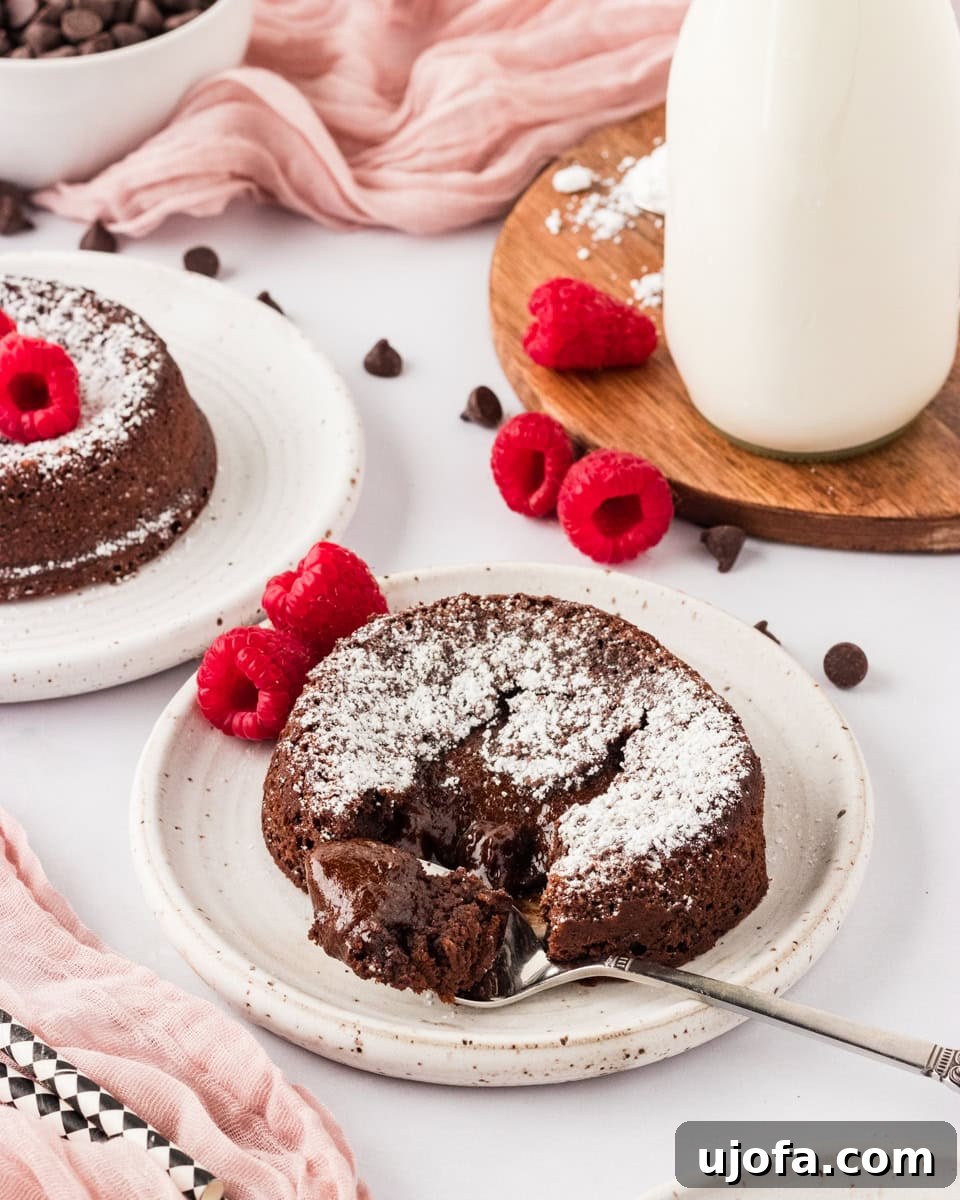 Molten Gluten Free Chocolate Bliss 2 A gluten-free chocolate lava cake on a white plate, dusted with powdered sugar, surrounded by fresh raspberries and chocolate chips. A fork reveals the gooey chocolate center. A pink cloth and a bottle of milk in the background add a soft touch.