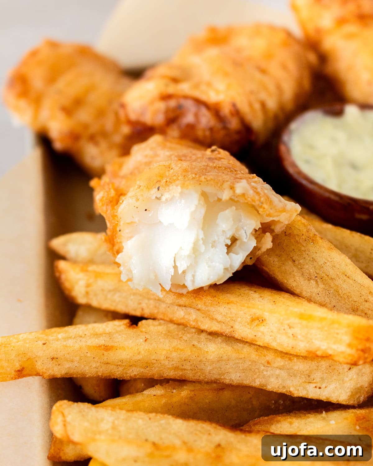 Close-up of a flaky, golden-brown piece of battered gluten-free fish with a bite taken out, resting on a bed of thick-cut fries, accompanied by a small bowl of dipping sauce.