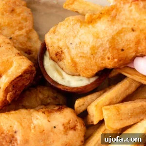 Crispy battered gluten-free fish being dipped into tartar sauce, surrounded by golden fries on a parchment-lined tray