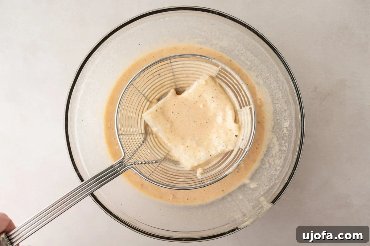 fish being dipped into a thick beige gluten-free batter, held in a wire mesh strainer above a glass mixing bowl containing more of the batter.