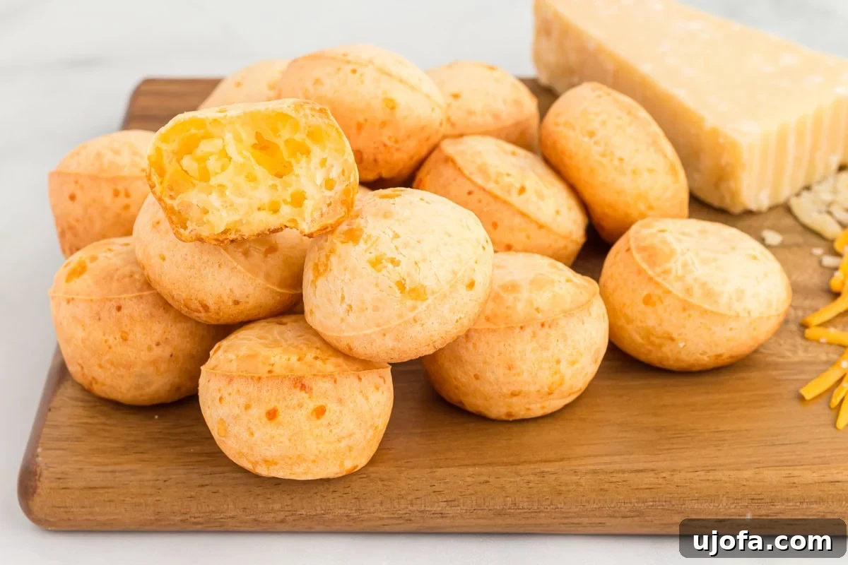 A stack of Brazilian cheese breads (pão de queijo) on a wooden cutting board, with one bread broken open to reveal its soft, cheesy, and airy interior. Grated cheese and a block of cheese are also visible in the background.