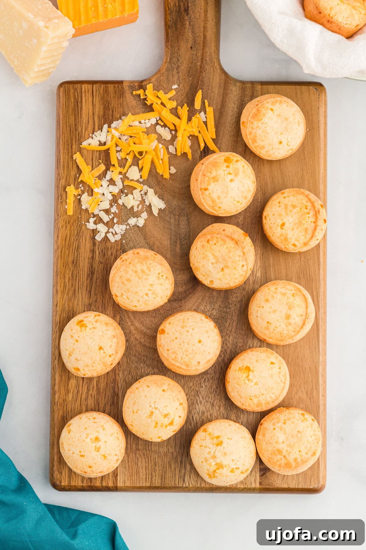 A wooden cutting board displaying golden-brown Pão de Queijo (Brazilian cheese bread), with shredded cheddar and Parmesan cheese arranged decoratively on the board. Blocks of cheese are visible in the corner, along with a basket of bread wrapped in a white cloth. A teal napkin adds a pop of color to the composition.