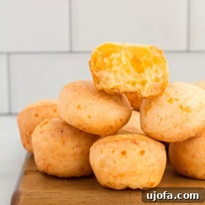 A stack of golden-brown Pão de Queijo (Brazilian cheese bread) on a wooden board, with one piece on top cut open to reveal its airy, light, and cheesy interior.