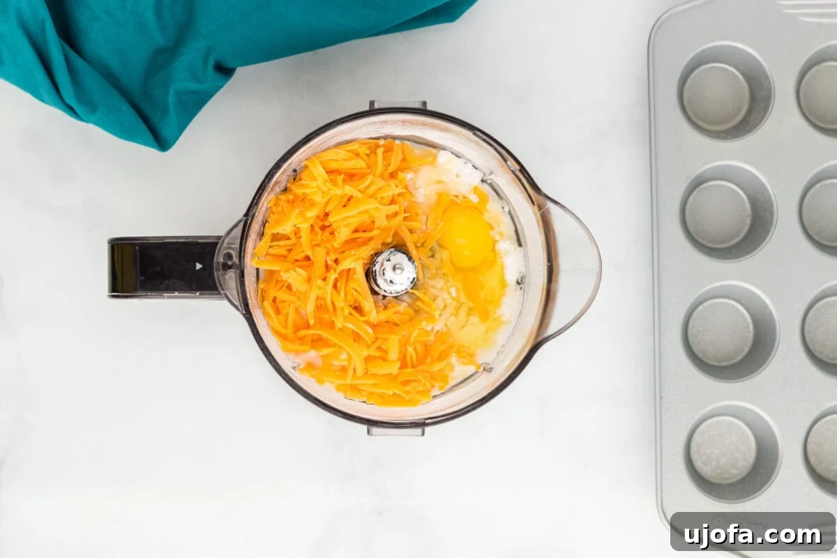 A food processor bowl filled with ingredients, including shredded cheddar cheese, eggs, and other components, ready to be blended. A muffin tin is placed in the background on a white surface, with a teal cloth nearby.