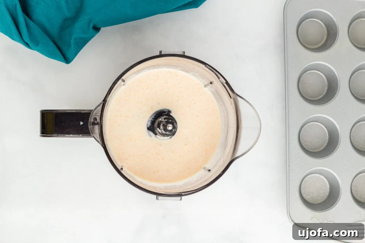 A food processor bowl containing a smooth, blended mixture of ingredients with a creamy texture. A muffin tin is placed in the background on a white surface, with a teal cloth nearby.