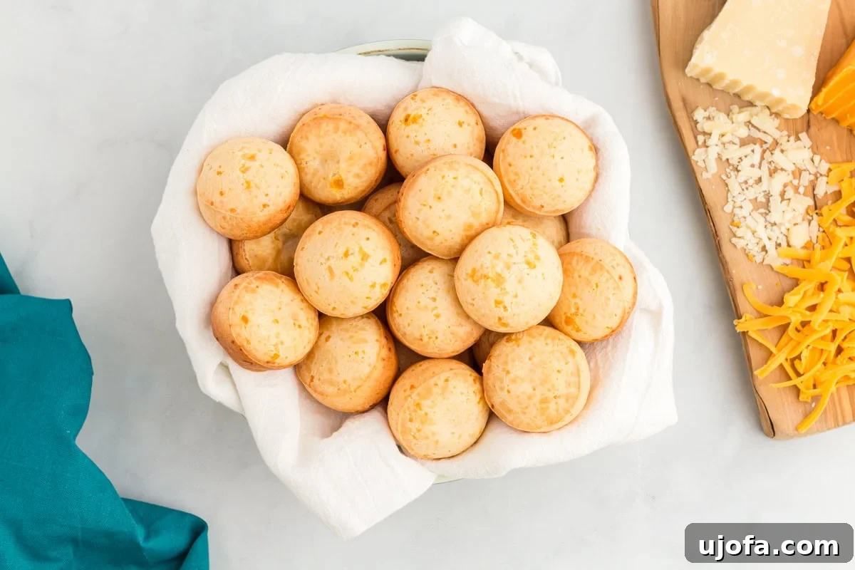 A basket lined with white cloth holds several round, golden-brown Brazilian cheese breads (pão de queijo). A wooden board with grated cheese and cheese blocks is partially visible in the background.