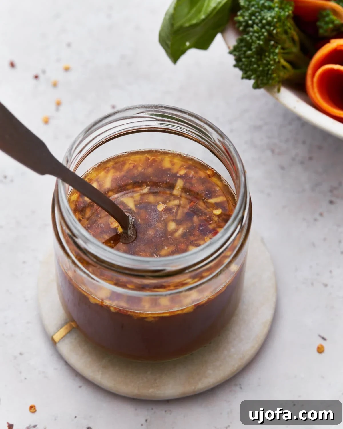 Delicious Gluten-Free Stir Fry Enhancer 2 A close-up of a jar filled with gluten-free stir-fry sauce, featuring visible bits of garlic and red chili flakes. The jar sits on a small round coaster, with vibrant fresh broccoli and carrots blurred in the background.