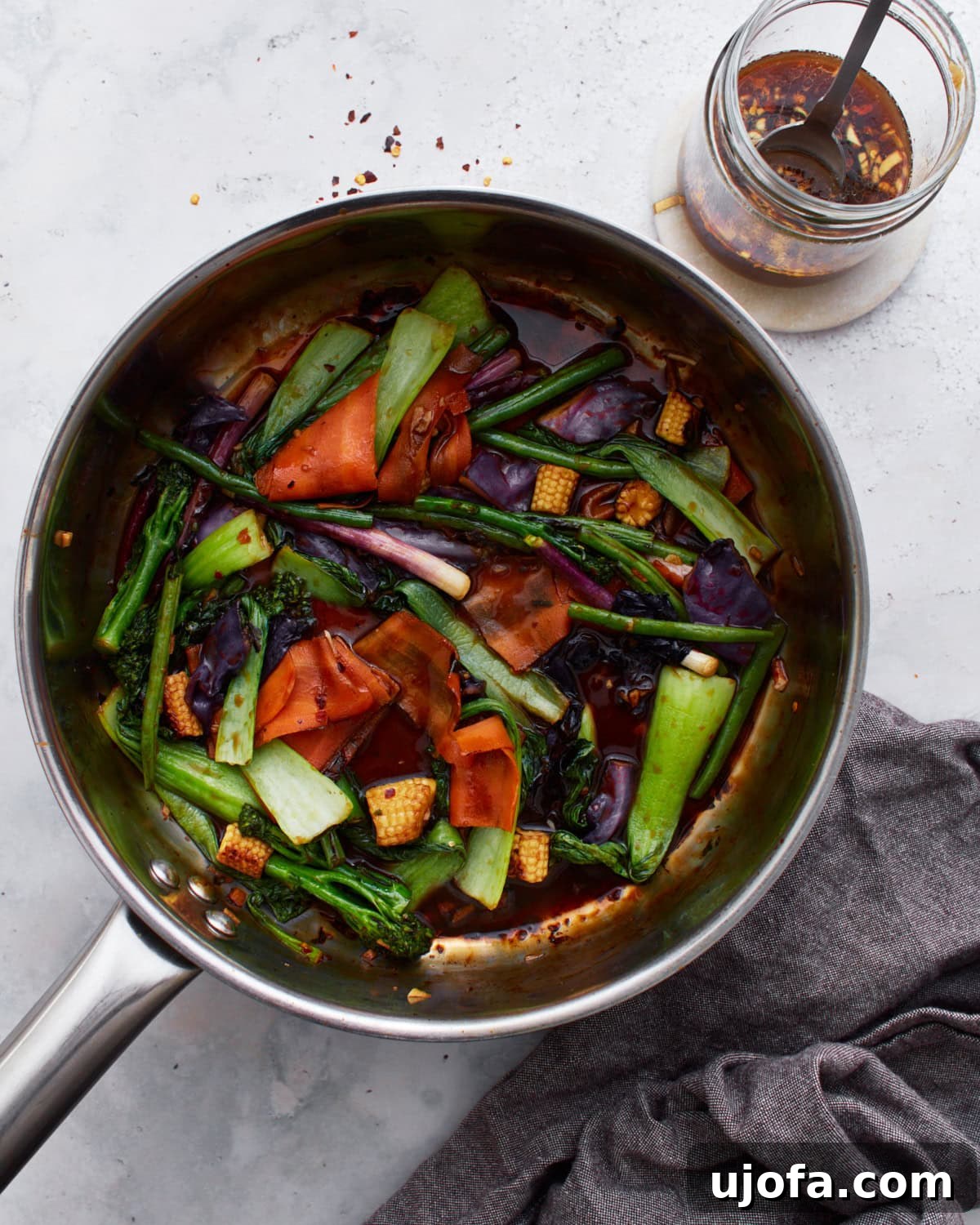 Delicious Gluten-Free Stir Fry Enhancer 10 A sauté pan filled with colorful stir-fried vegetables, including bok choy, baby corn, carrots, and purple cabbage, tossed in a glossy gluten-free stir-fry sauce. A jar of the sauce sits nearby.