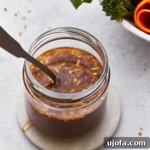 A close-up of a jar filled with gluten-free stir-fry sauce, featuring visible bits of garlic and red chili flakes. The jar sits on a small round coaster, with vibrant fresh broccoli and carrots blurred in the background.