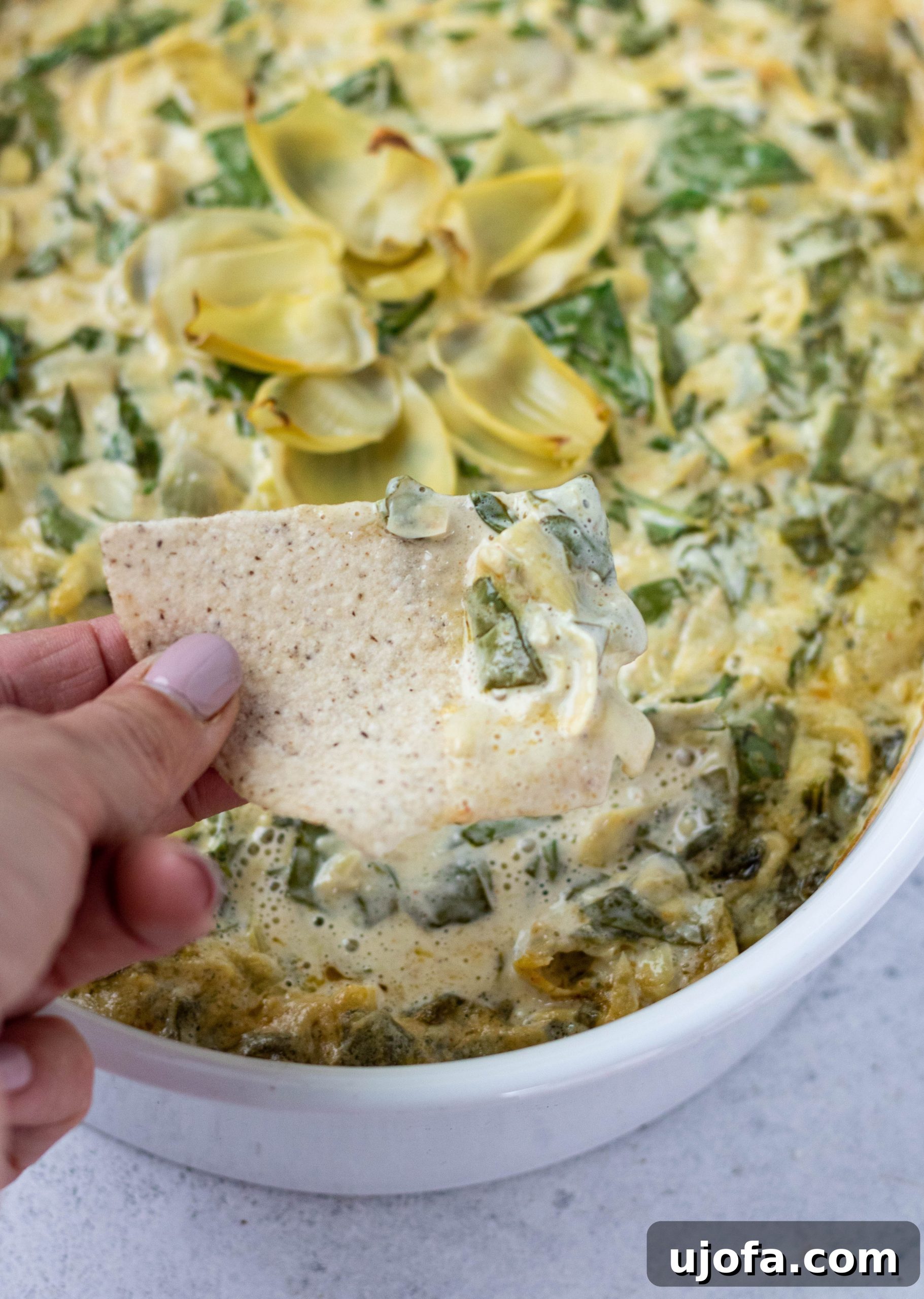 A hand expertly scooping a generous portion of creamy dairy-free spinach artichoke dip with a tortilla chip from a casserole dish.