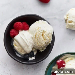 A black bowl filled with scoops of creamy white keto vanilla ice cream, garnished with fresh raspberries and blueberries. A single strawberry sits nearby on the surface.