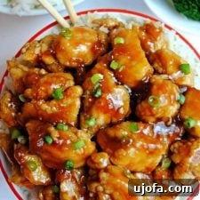A close-up shot of a bowl filled with delectable gluten-free apricot chicken, garnished and ready to be served, with chopsticks, broccoli, and rice blurred in the background.