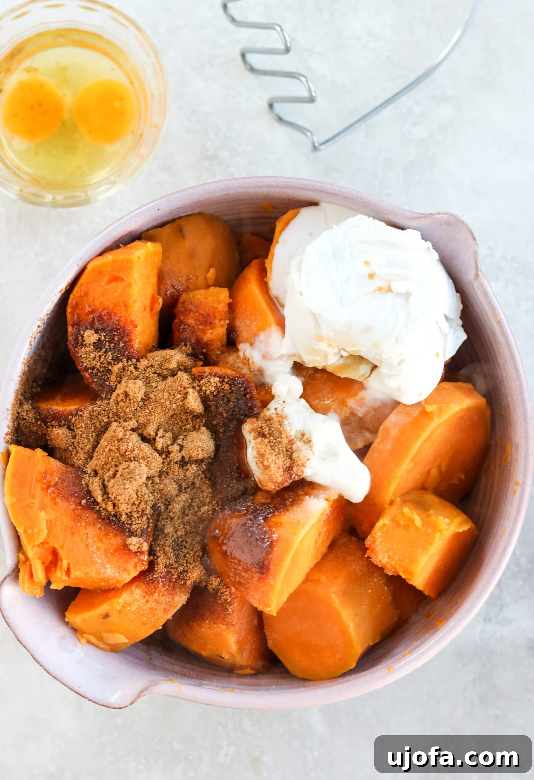 Mashed sweet potatoes in a large bowl with various ingredients around it, including coconut sugar and an egg, ready to be combined for the casserole base.
