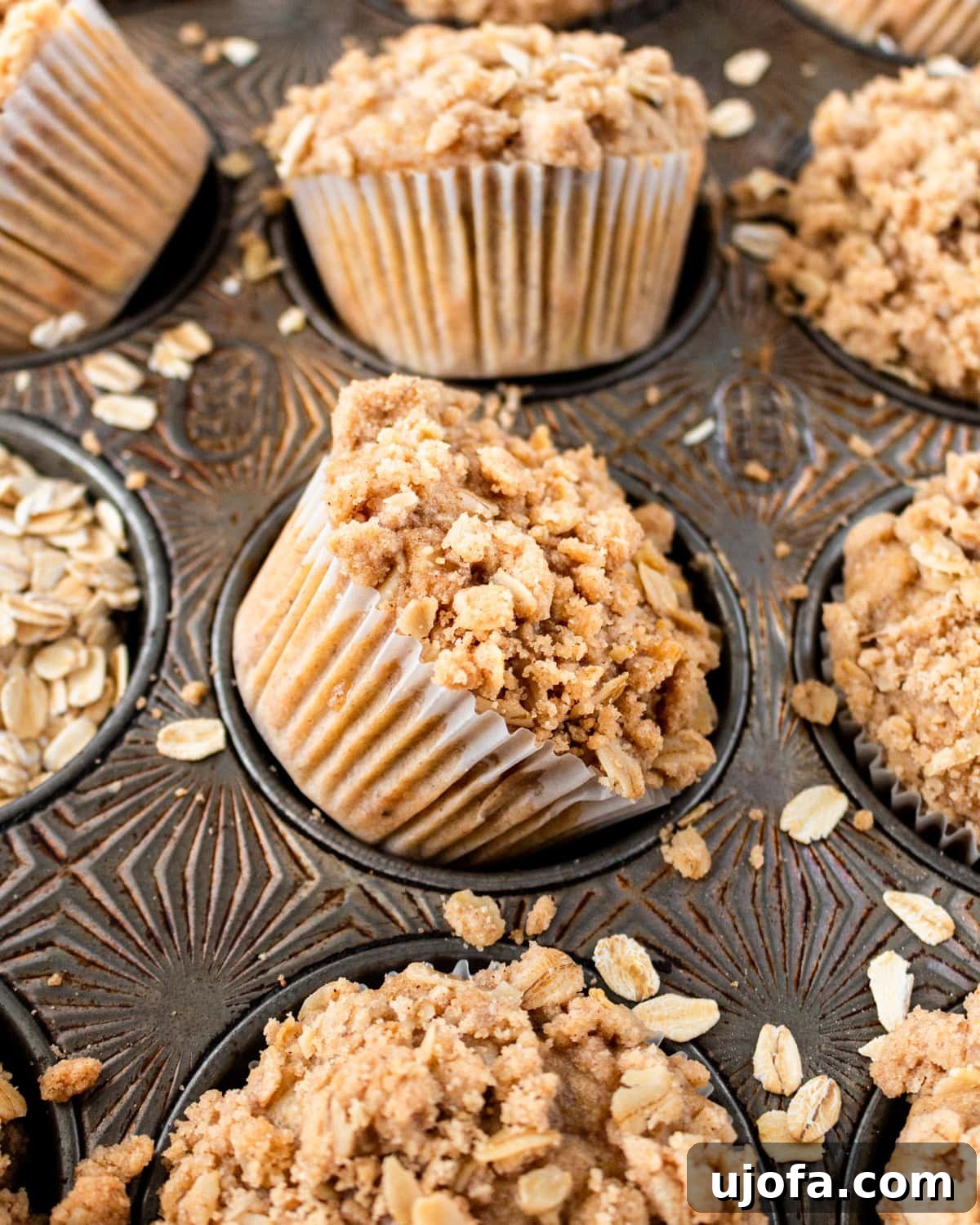 Close-up of banana oatmeal muffins in a rustic muffin tin, topped with a crumbly oat streusel and surrounded by scattered oats.